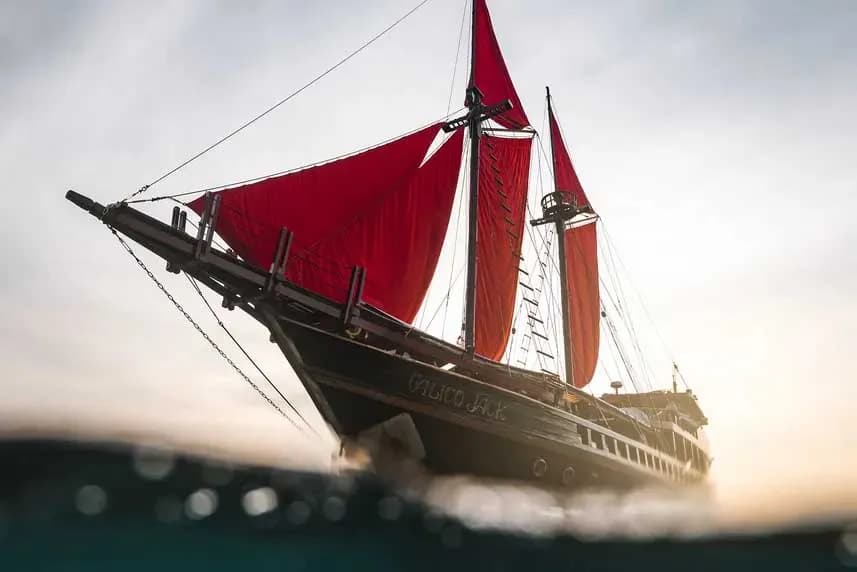 A red sailboat named Calico Jack glides across the ocean with a vibrant sunset illuminating the sky behind it. A red sailboat named Calico Jack glides across the ocean with a vibrant sunset illuminating the sky behind it.